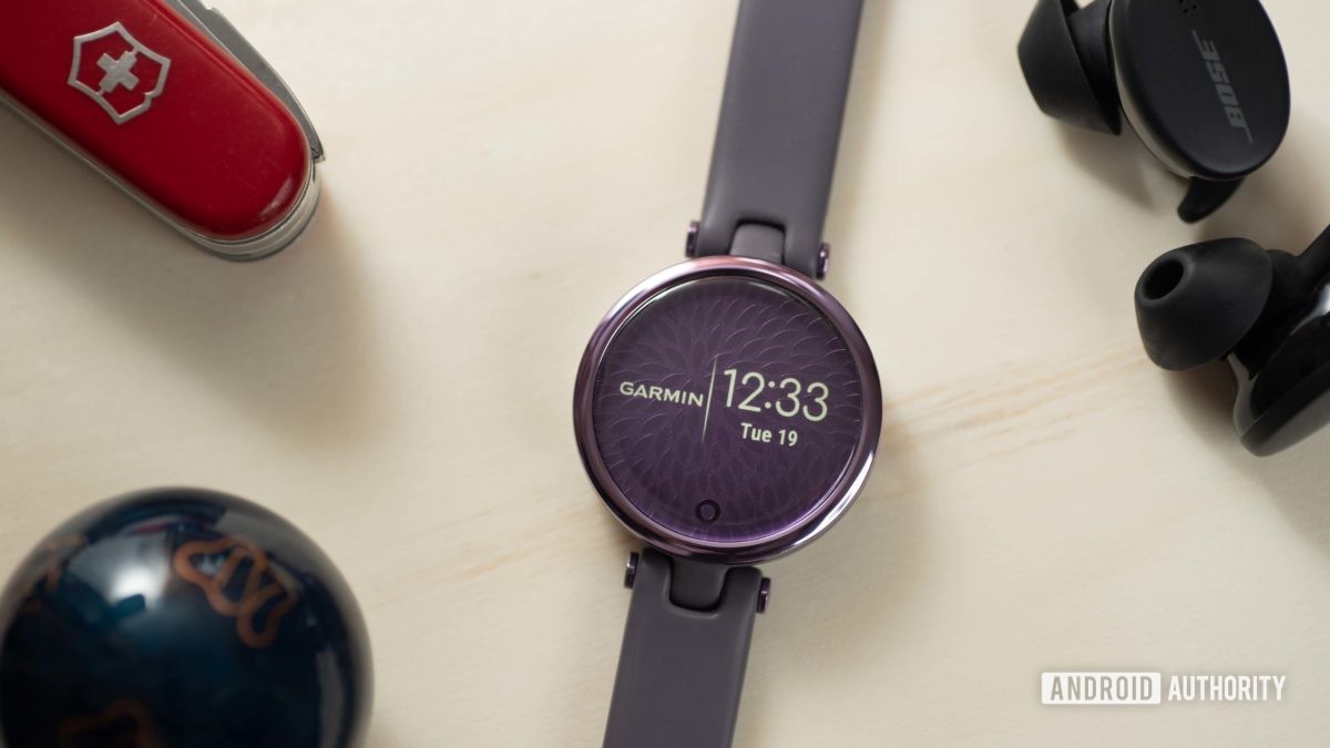 Het Garmin Lily Sport Edition smartwatch geeft de tijd en het Garmin-logo weer en rust naast een paar Bose Sport Earbuds en een Zwitsers zakmes.