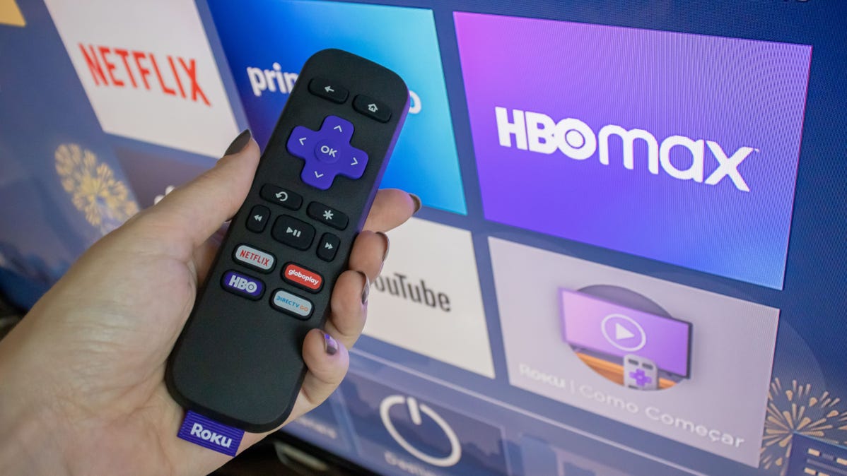 Roku remote in hand in front of a Roku home screen.