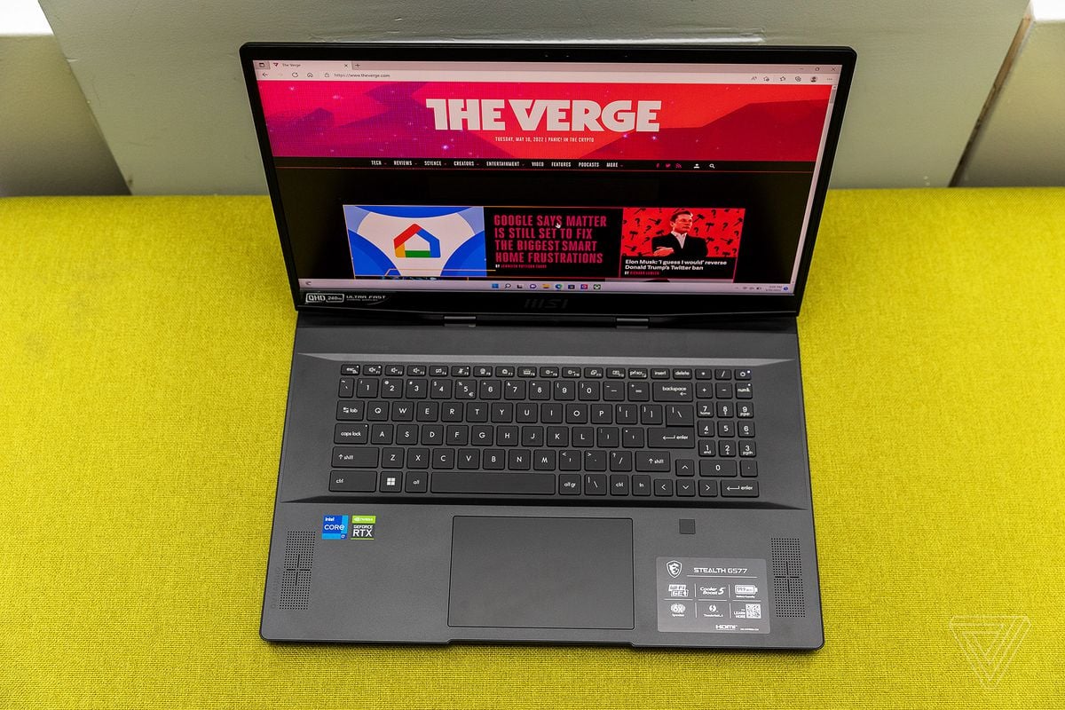 The MSI GS77 Stealth keyboard deck seen from above on a yellow fabric bench. The screen displays The Verge homepage.