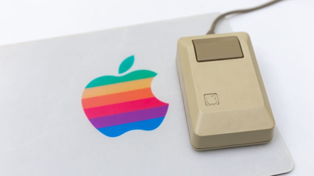A vintage Apple computer mouse on a mousepad with the rainbow Apple logo.