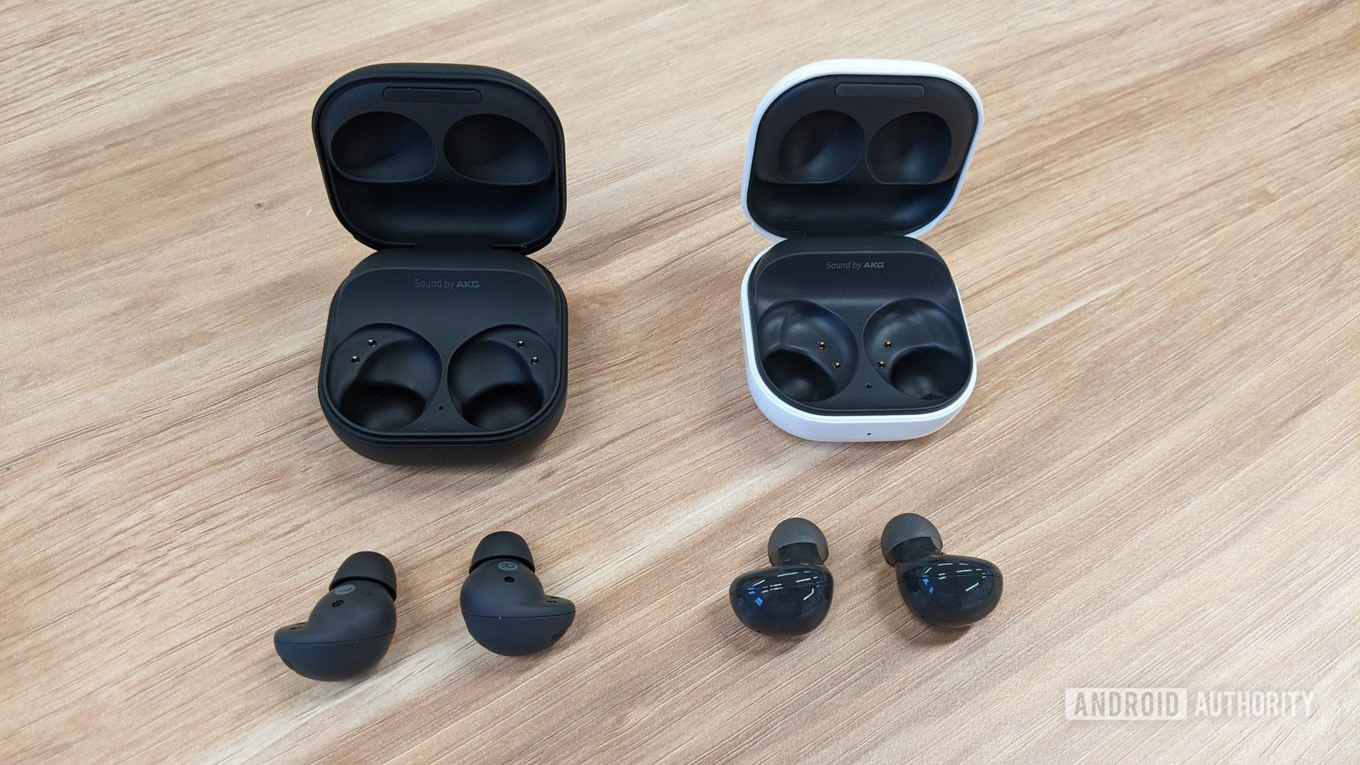 The Samsung Galaxy Buds 2 Pro in graphite sitting outside of their case to the left of the Galaxy Buds 2 in graphite outside of their (white) case on a table.