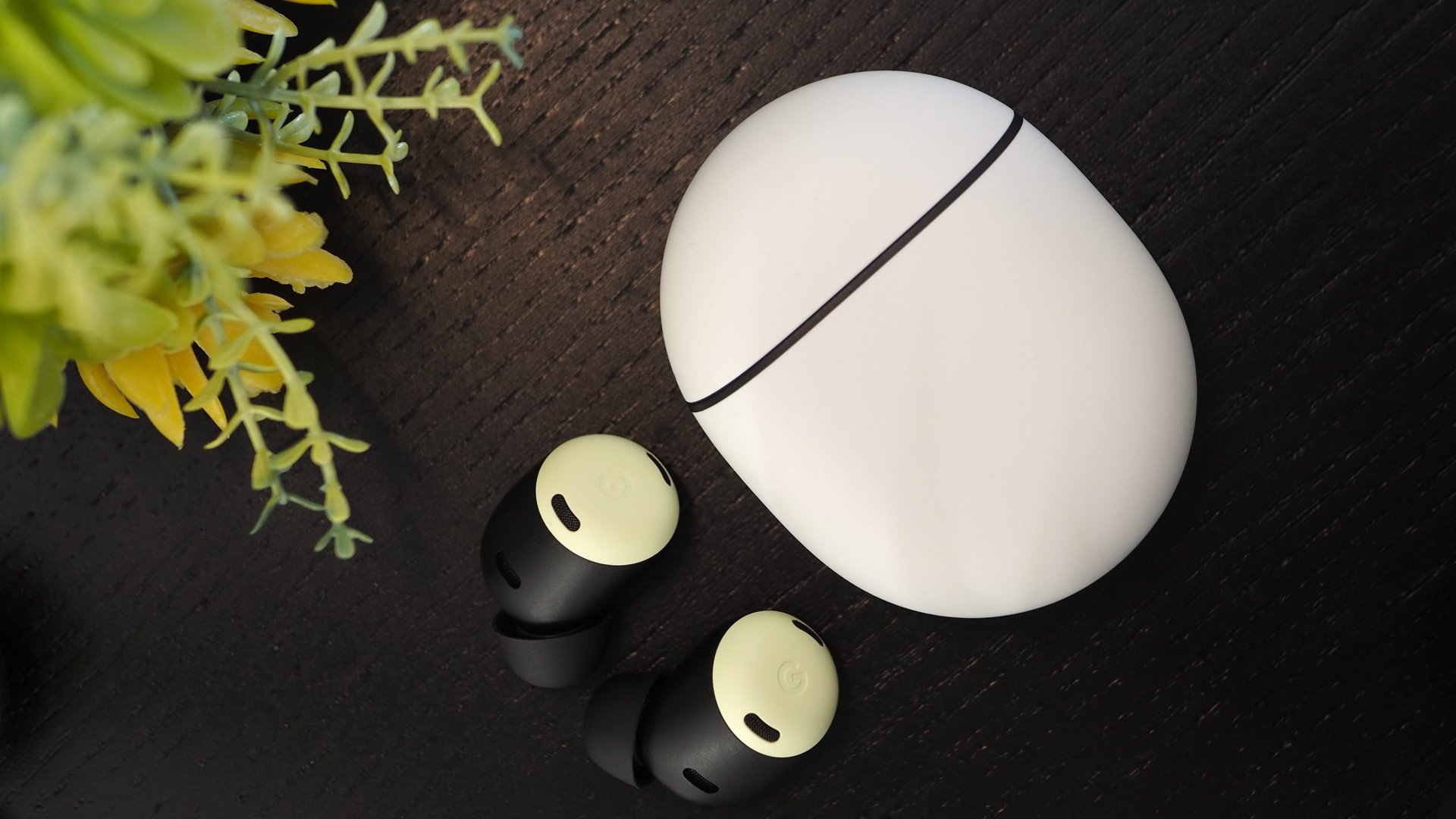 The Pixel Buds Pro outside ofo their case sittin on a dark wood table next a plastic plant.